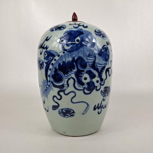 Puede incluir: Un jarrón de cerámica azul y blanco con tapa. El jarrón presenta una ilustración detallada de una criatura mítica en azul sobre un fondo blanco. La tapa está rematada con un adorno marrón oscuro.