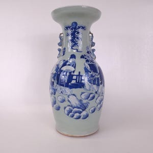 Peut inclure: Un vase en céramique vert clair avec un bord évasé et deux petites poignées. Le vase est décoré d'illustrations bleues et blanches de personnages et de motifs floraux. Le vase a un corps bulbeux et une surface légèrement texturée.