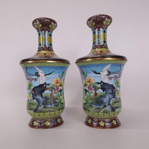 Pode incluir: Um par de vasos cloisonné a condizer com um fundo de céu azul vibrante, grous brancos em voo, motivos florais coloridos e formações rochosas escuras. Os vasos têm um pescoço alargado, um corpo largo e uma base circular com detalhes em metal.