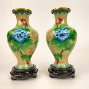 May include: A pair of cloisonné vases with floral designs. The vases are gold with blue, green, and red flowers. They are on black wooden stands.