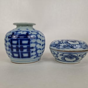 May include: A two-piece blue and white ceramic set. The set includes a small vase with a double happiness symbol and a round lidded box with floral designs. The pieces are painted in a traditional Chinese style.