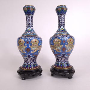 Pode incluir: Um par de vasos cloisonné a condizer com um fundo azul vibrante, padrões florais multicoloridos intrincados e figuras de leões estilizadas. Cada vaso tem um pescoço fino, um corpo bulboso e repousa sobre uma base de madeira escura esculpida.