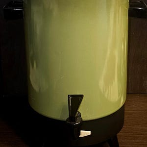 May include: A vintage green and black electric coffee urn. The urn has a cylindrical body with a lid and a spigot. It sits on three black legs and has black handles on the sides. The lid and handle are black.