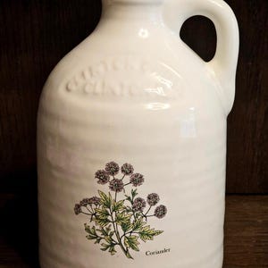 May include: White ceramic jug with a handle, featuring a botanical illustration of coriander. The jug has embossed text near the top. The coriander illustration includes green leaves and purple flower clusters. The jug is on a dark wooden surface.