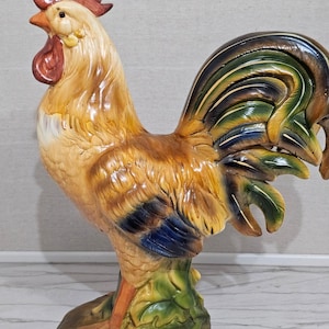 May include: A ceramic rooster figurine featuring a glossy finish with a vibrant color palette of tan, brown, dark blue, and green. The rooster stands on a sculpted base with detailed feathers, a prominent red comb, and a red wattle on its neck.