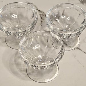 May include: Three clear, stemmed dessert dishes with a diamond-cut pattern. The bowls are round with a wide base and a delicate stem. The dishes are arranged on a light-colored surface, showcasing their transparency and design.