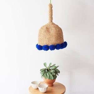 May include: A woven beige pendant light with a bell shape, adorned with a row of vibrant blue pom-poms along the bottom edge. The light hangs from a green cord. A small potted plant and two white bowls sit on a wooden table below.