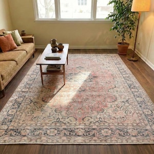 Op de afbeelding: Een groot, rechthoekig vloerkleed met een vervaagde, vintage uitstraling. Het vloerkleed heeft een centraal ontwerp in roze en crèmetinten, omrand door een donkerblauw en crèmekleurig bloemenpatroon. Het ligt op een donkere houten vloer.