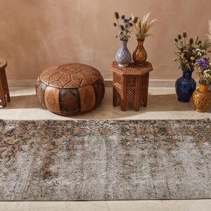 Puede incluir: Escena de diseño de interiores con un otomán de cuero marrón, una alfombra estampada, mesas de madera tallada y jarrones decorativos con flores secas. La paleta de colores incluye marrones, beiges y azules, creando un ambiente cálido.