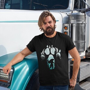 Op de afbeelding: Zwart t-shirt met een groot, verweerd pootafdrukontwerp in wit en blauwgroen. Binnen de pootafdruk bevindt zich een silhouet van een beer en bomen. Het t-shirt wordt door een persoon gedragen.