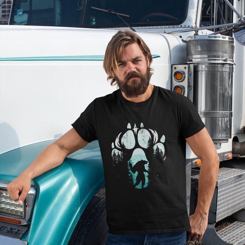 Op de afbeelding: Zwart t-shirt met een groot, verweerd pootafdrukontwerp in wit en blauwgroen. Binnen de pootafdruk bevindt zich een silhouet van een beer en bomen. Het t-shirt wordt door een persoon gedragen.