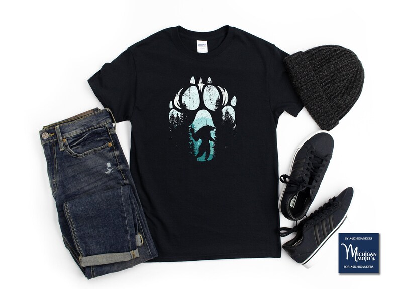 Op de afbeelding: Zwart t-shirt met een Bigfoot-afbeelding in een berenpootafdruk. Het ontwerp toont een silhouet van Bigfoot in een boslandschap, met een zwarte muts, donkerblauwe jeans en zwarte sneakers. Het shirt is van Michigan Mojo.