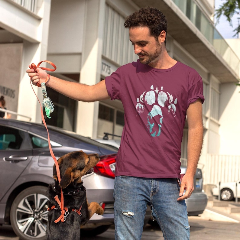 Op de afbeelding: Bordeauxrode crewneck t-shirt met een witte en blauwgroene berenpootafdruk. Het shirt wordt gecombineerd met een blauwe jeans. Een hond draagt een oranje harnas en een riem.