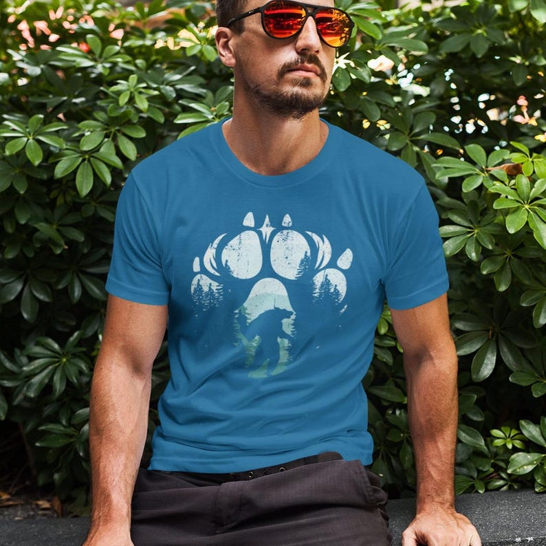 Op de afbeelding: Een tealkleurig t-shirt met een wit berenpootafdrukontwerp met een berensilhouet en bomen. Dit grafische t-shirt is gemaakt van een zacht materiaal en is geschikt voor vrijetijdskleding.