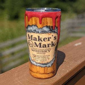 May include: A red and white insulated tumbler with a Maker's Mark whiskey label. The tumbler is decorated with a wood grain pattern and a red drip effect.