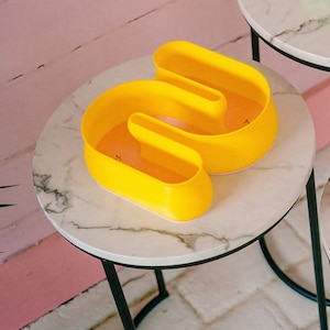 May include: A yellow, three-dimensional, abstract sculpture shaped like the letter "M" sits on a white marble table with black legs.
