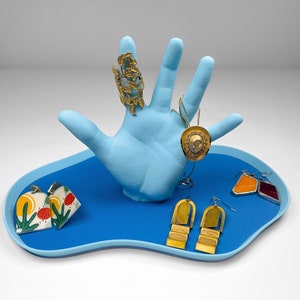 A blue hand with fingers extended on top of a blue blob shaped tray against a plain white background. The hand holds a ring, a necklace and a few pairs of earrings.