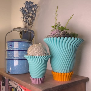May include: Two turquoise ceramic planters with a wavy design. The larger planter has a bright orange base and holds a variety of succulents. The smaller planter has a purple base and holds a small cactus.