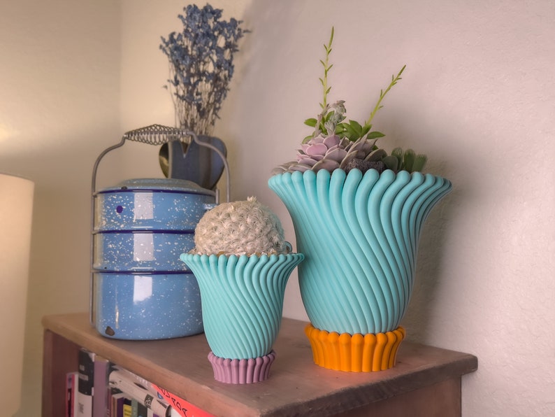 May include: Two turquoise ceramic planters with a wavy design. The larger planter has a bright orange base and holds a variety of succulents. The smaller planter has a purple base and holds a small cactus.