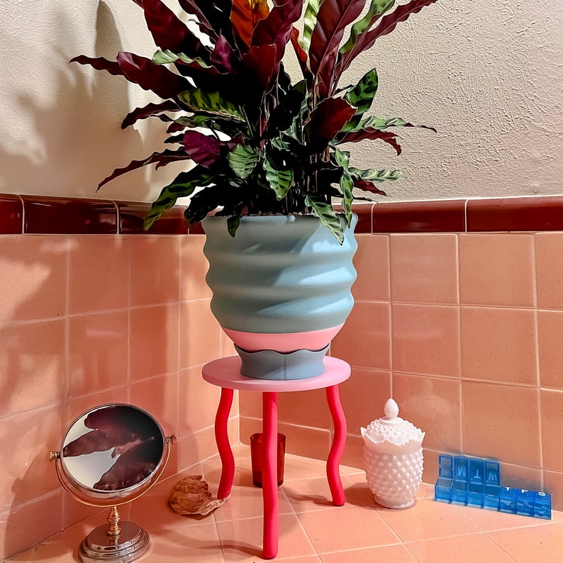 Puede incluir: Una maceta de cer&aacute;mica rosa y azul con un dise&ntilde;o ondulado se asienta sobre un soporte de madera rosa y blanco. Una planta verde frondosa con hojas rojas y moradas crece en la maceta. La planta est&aacute; en un ba&ntilde;o con azulejos rosa y blancos.