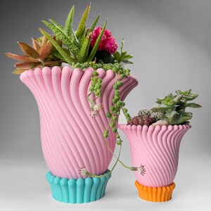 May include: Two pink ceramic planters with a wavy, ribbed design. The larger planter has a turquoise base and the smaller planter has an orange base. Both planters are filled with succulents and other greenery.