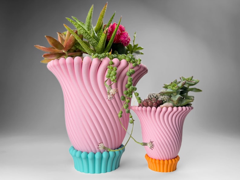 May include: Two pink ceramic planters with a wavy, ribbed design. The larger planter has a turquoise base and the smaller planter has an orange base. Both planters are filled with succulents and other greenery.