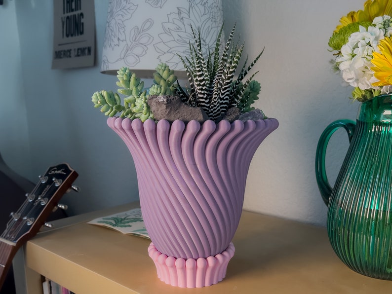 May include: A purple ceramic planter with a unique swirl design and a pink base. The planter is filled with a variety of succulents, including a zebra plant.