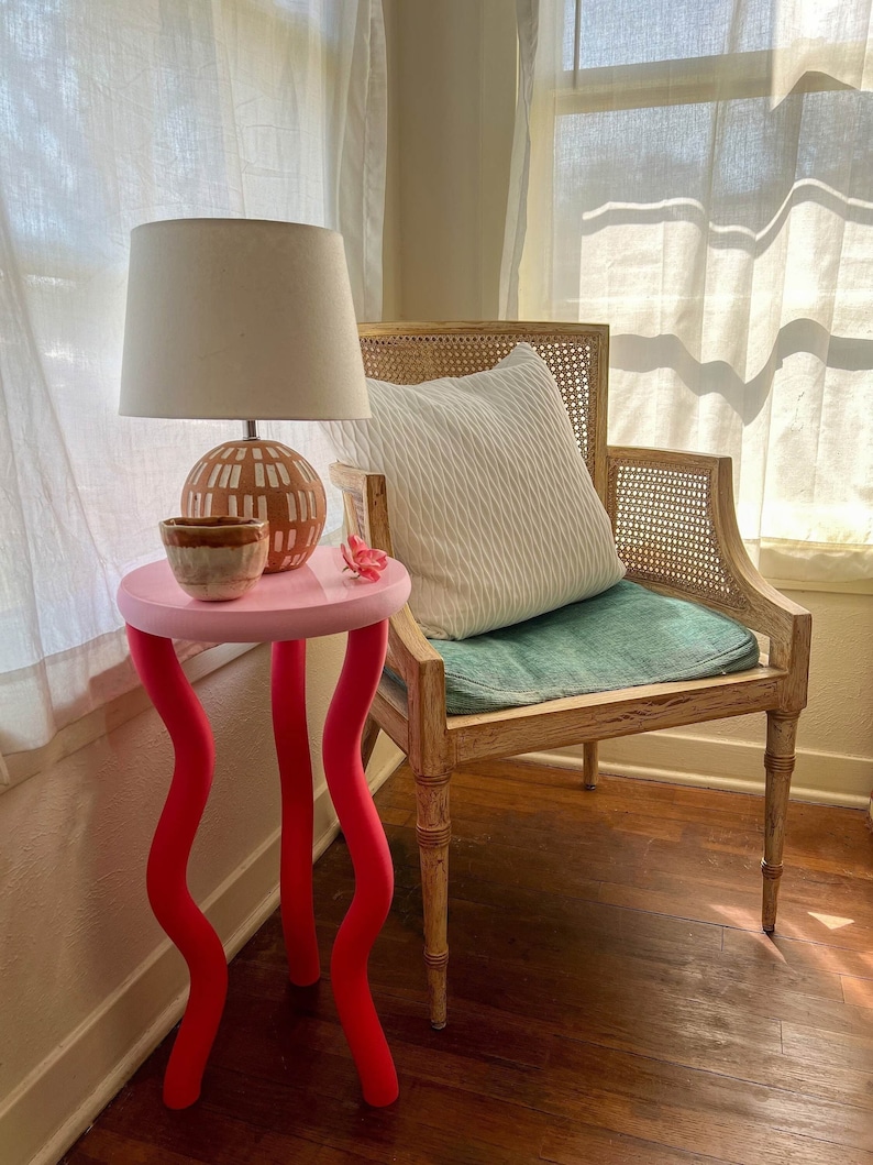 Red and Pink Side Table With Wavy Wiggly Legs holds a small lamp and flower and sits next to a vintage chair in a naturally lit lovely room