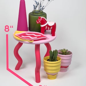 Puede incluir: Una mesa cuadrada rosa de 20 cm de lado con patas onduladas. La mesa tiene un plato rosa y blanco, un jarr&oacute;n verde con flores secas, un caballo de madera rojo y dos peque&ntilde;as macetas.