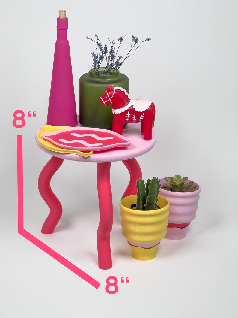 Puede incluir: Una mesa cuadrada rosa de 20 cm de lado con patas onduladas. La mesa tiene un plato rosa y blanco, un jarr&oacute;n verde con flores secas, un caballo de madera rojo y dos peque&ntilde;as macetas.