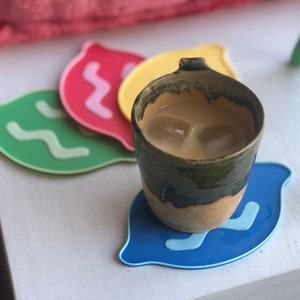 Puede incluir: Una taza de cerámica con un esmalte verde y un borde marrón, llena de café helado y cubitos de hielo, sentada sobre un posavasos azul, amarillo, verde y rojo con un diseño ondulado.