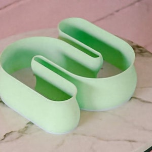 May include: A light green ceramic planter shaped like the letter 'W' with a smooth, rounded surface. The planter is sitting on a white marble surface.