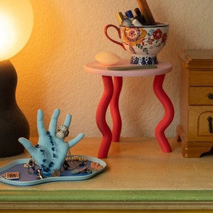 Puede incluir: Un porta anillos de cer&aacute;mica azul en forma de mano con un anillo blanco en el dedo &iacute;ndice. La mano est&aacute; sobre una bandeja azul con otras joyas. Una peque&ntilde;a mesa rosa con patas rojas sostiene una taza de cer&aacute;mica blanca con dise&ntilde;os florales coloridos y un peine. La mesa est&aacute; al lado de un armario de madera.