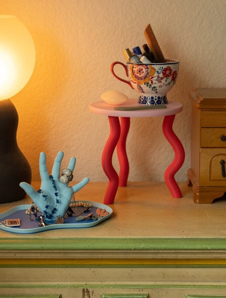 Puede incluir: Un porta anillos de cer&aacute;mica azul en forma de mano con un anillo blanco en el dedo &iacute;ndice. La mano est&aacute; sobre una bandeja azul con otras joyas. Una peque&ntilde;a mesa rosa con patas rojas sostiene una taza de cer&aacute;mica blanca con dise&ntilde;os florales coloridos y un peine. La mesa est&aacute; al lado de un armario de madera.