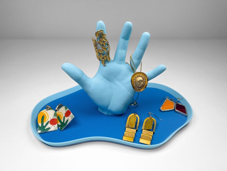 A blue hand with fingers extended on top of a blue blob shaped tray against a plain white background. The hand holds a ring, a necklace and a few pairs of earrings.