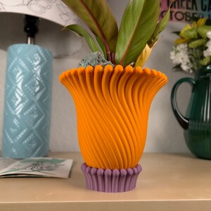 May include: A bright orange, 3D printed flower pot with a swirling design. The pot has a purple base and is sitting on a light brown surface.
