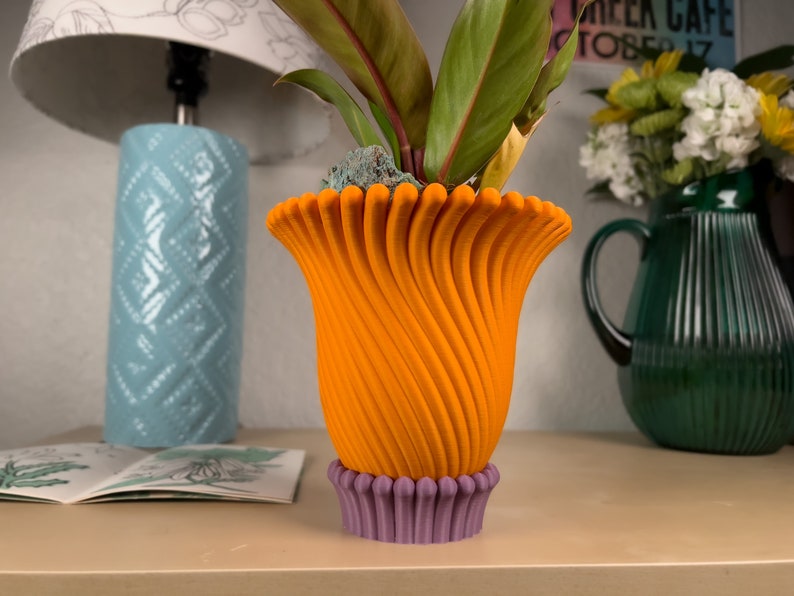 May include: A bright orange, 3D printed flower pot with a swirling design. The pot has a purple base and is sitting on a light brown surface.