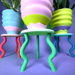 Puede incluir: Tres macetas de cer&aacute;mica de colores con dise&ntilde;os ondulados, cada una sentada en un soporte verde con patas onduladas. Las macetas son azul claro, rosa y verde con acentos amarillos.