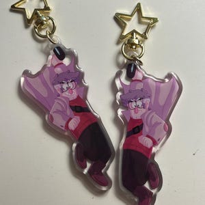 以下が含まれることがあります： メガネ、赤いセーター、紫のマントを着た漫画のキャラクターが描かれた、透明なアクリルキーホルダー2つ。各キーホルダーには、金色の星型の留め金が付いています。キャラクターは厳しい表情をしています。