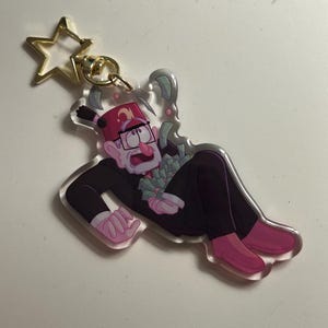 以下が含まれることがあります： 黒いスーツ、ピンクの靴、赤いフェズを身に着けた漫画のキャラクターが描かれた、透明なアクリルキーホルダー。キャラクターはお金の山を持って横たわっています。キーホルダーには金色の星型の留め金が付いています。