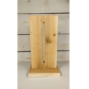 Può includere: Una lampada da tavolo in legno con finitura in legno naturale. La lampada ha una base rettangolare e un paralume rettangolare alto con una striscia verticale di luci LED al centro.