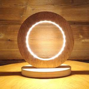 May include: A wooden table lamp with a circular base and a round, light-up ring in the centre. The lamp is lit and casting a warm glow.