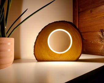 May include: A wooden lamp with a circular light in the center. The lamp is made from a cross-section of a tree trunk, showcasing the natural wood grain and bark. The light emits a warm glow, creating a cozy ambiance. A potted plant is visible in the background.