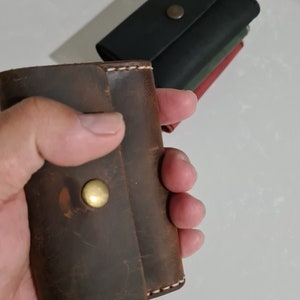 May include: A brown leather key holder with a brass button closure is held in a hand. The key holder has white stitching and a distressed finish. Other key holders in black, green, and red are in the background.