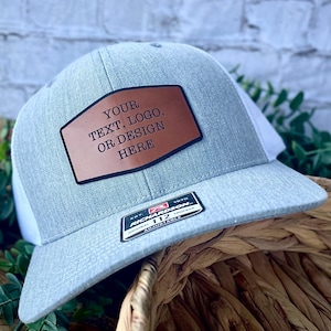 May include: A gray trucker hat with a white mesh back and a brown leather patch. The patch has the text "YOUR TEXT, LOGO, OR DESIGN HERE". The hat is adjustable.