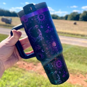 Può includere: Una tazza da viaggio in acciaio inossidabile nero con una finitura iridescente viola e nera. La tazza presenta un design celeste con lune, stelle e raggi di sole. La tazza ha un coperchio con manico.