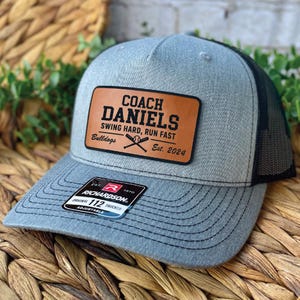 May include: A grey and black baseball cap with a mesh back. The front has a brown leather patch with the text "COACH DANIELS SWING HARD, RUN FAST Bulldogs Est. 2024". The cap has a curved peak and an adjustable strap.
