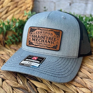 May include: A grey trucker hat with a black mesh back. The hat features a brown leather patch with the words "CERTIFIED SHADETREE MECHANIC" in a decorative font. The hat has a curved brim and an adjustable strap.
