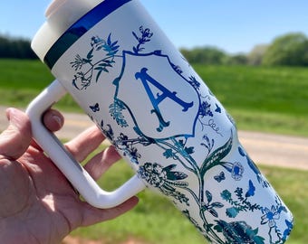 May include: White stainless steel travel mug with a blue and white floral design and the letter "A" in a blue and white shield. The mug has a white handle.