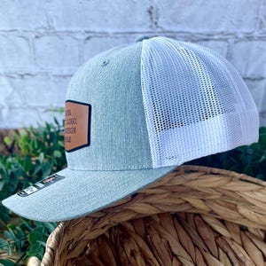 Side profile of a custom Richardson 112 trucker hat in heather grey with white mesh and engraved leather patch on the front.